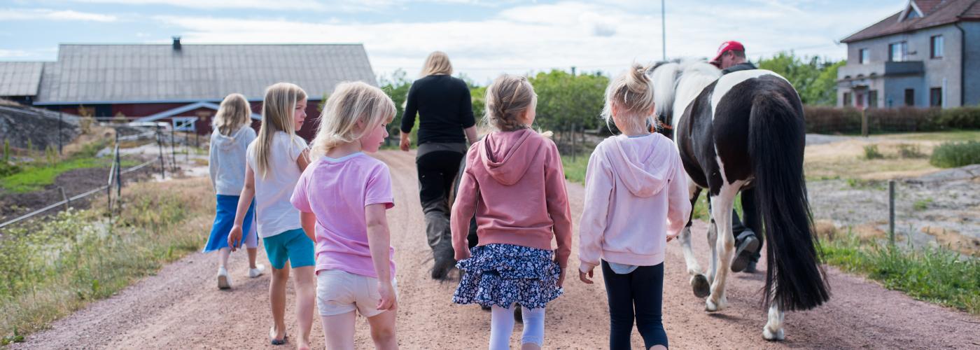 Fem barn, två vuxna och en häst går på en grusväg, med ryggen till, bort från kameran. 