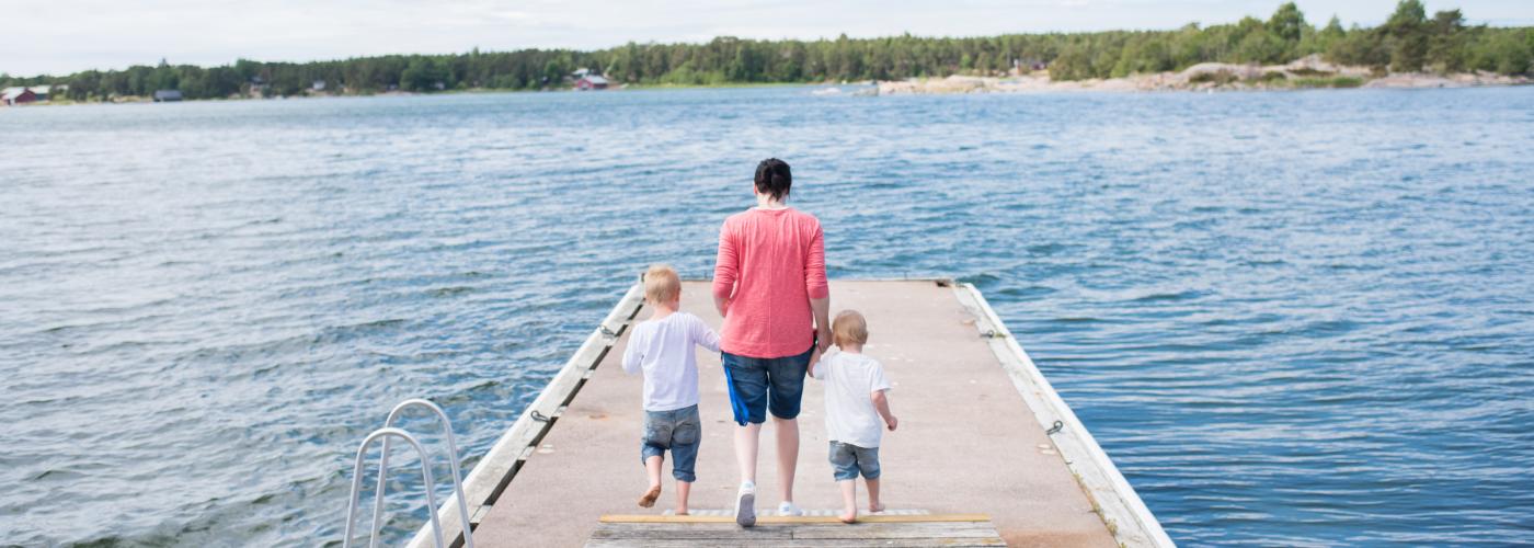 En kvinna med ett varsitt barn i handen går, bort från kameran, ut på en brygga. Blå himmel och sommarsolen skiner.